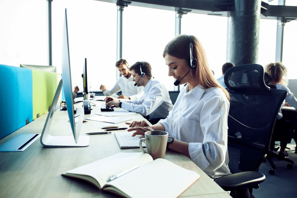 A woman wearing a headset types on her computer while sitting at a desk.